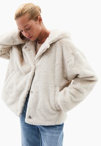 Kurtka z futra ekologicznego z kapturem w jasnym beżu o miękkiej fakturze, z dużymi kieszeniami z przodu i zapięciem na napy. Nosi ją osoba na beżowej bluzce w połączeniu z niebieskimi dżinsami.