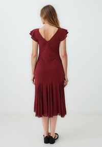Bordeaux rode jurk met een V-rug, korte gerimpelde mouwen en een uitlopende rok. Gemaakt van soepel materiaal, met een vloeiende silhouette.