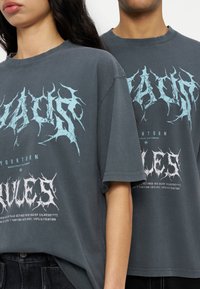 Δυο άτομα φορούν υπερμεγέθη σκούρου γκρι μπλουζάκια με σχέδια από γραφικό κείμενο σε αιχμηρά, αραιωμένα μπλε και λευκά στοιχεία.