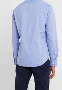 Světlomodrá košile s dlouhými rukávy, jednobarevná, zakřivený lem, manžety na knoflíky a klasický límec. Nošena s tmavými kalhotami.