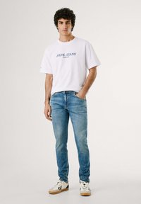 Balts kokvilnas T-krekls ar zilu "Pepe Jeans London" logotipu, kombinēts ar gaiši ziliem džinsiem un baltiem kedām ar melniem akcentiem.