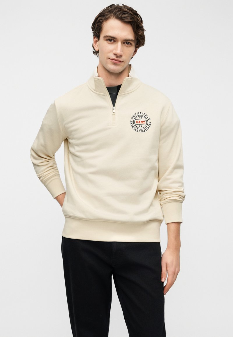 Felpa beige con zip e maniche lunghe, caratterizzata da un logo circolare sul petto. Tessuto morbido con orlo e polsini a coste.