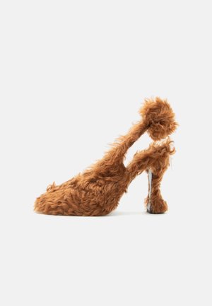 Brown high-heeled shoe covered in shaggy fur with a fur pom-pom decoration on the ankle strap against a white background.