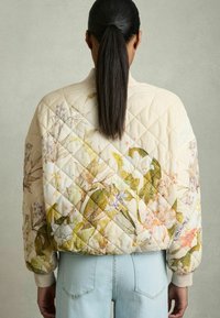 Veste bomber matelassée avec des motifs floraux dans des teintes pastel douces, dotée de poignets côtelés et d'une coupe décontractée, associée à un jean bleu clair.