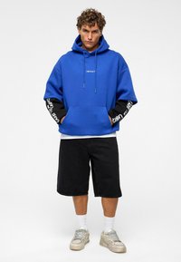 Blauwe hoodie met zwarte mouwen, voorzien van een wit "IMPACT" logo. Draag met zwarte shorts en grijze sneakers. Witte sokken zichtbaar.