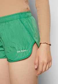 Calções verdes, leves, com um cós elástico, bolsos laterais e uma bainha curva. Apresentam texto branco bordado na parte da frente.