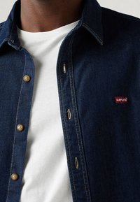 Camicia di jeans blu scuro con colletto strutturato, bottoni in ottone e patch con logo rosso Levi's. Maglietta bianca visibile sotto.