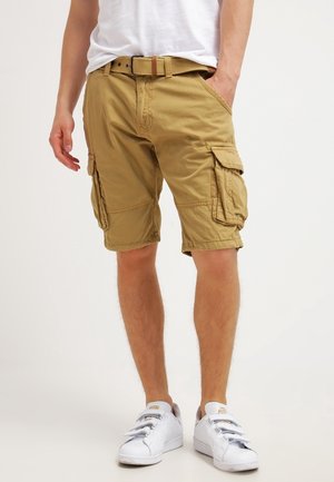 Homme portant un short cargo beige avec plusieurs poches, une ceinture assortie, des baskets blanches et un t-shirt blanc, debout avec une main dans la poche.