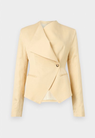 Μπεζ σακάκι με ασύμμετρο πέτο, κλείσιμο με ένα μπεζ κουμπί και μακριά μανίκια, εμφανιζόμενο σε ανοιχτό φόντο.