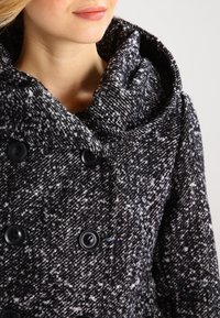 Svart och vit prickig kappa med huva, prydd med stora svarta knappar och en texturerad tyg. Närbild på halsringningen och knapparna.