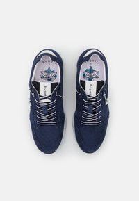Tummansiniset mokkanahka sneakersit, joissa on valkoiset nauhat, kumipohja ja koristeellinen sisäpohjan muotoilu, jossa on lootuskukka ja brändilogo.