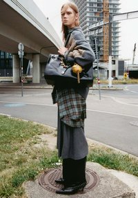 Schwarze Handtasche mit goldfarbenen Beschlägen und Charms, grauer oversized Pullover, kariertes Hemd darunter, langer grauer Rock und spitze schwarze High Heels.