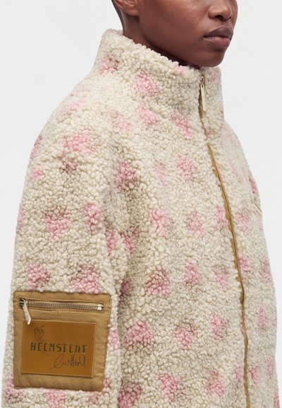 Personne portant une veste polaire texturée crème et rose avec un col montant et une poche zippée beige étiquetée Helmstedt sur la manche.