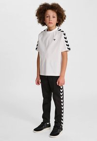 Hvid kortærmet t-shirt med sorte chevron-striber på skuldrene, kombineret med sorte træningsbukser med hvide sidelommer. Sorte sneakers.