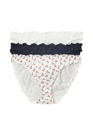 Jeune femme en camisole blanche et sous-vêtements à motif cerises, debout avec une main sur la hanche, sur un fond uni.