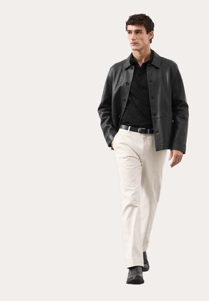 Schwarze Lederjacke mit Knopfverschluss, getragen über einem schwarzen Hemd und weißen Hosen. Das Modell steht vor neutralem Hintergrund und schaut geradeaus.