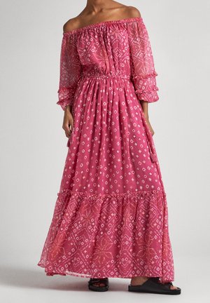 Woman wearing a pink off-shoulder maxi dress with geometric and floral patterns, long sheer sleeves, and black open-toe sandals.