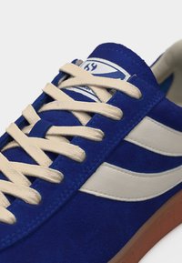 Blauer Wildledersneaker mit weißen Schnürsenkeln und Streifen, hellbrauner Sohle und Logo auf der Zunge, Nahaufnahme.