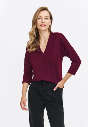 Bordowy pulower z dekoltem w serek i rękawami o długości trzy czwarte. Miękka tekstura, luźny krój, z centralnym detalem szwu. W połączeniu z czarnymi spodniami.