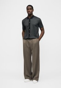 Mann trägt kurzärmeliges schwarzes Poloshirt mit Muster, weite beige Hose, weiße Turnschuhe, steht mit den Händen in den Taschen vor schlichte Hintergrund.