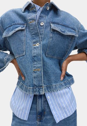 Persona vistiendo una chaqueta de mezclilla azul de capas sobre una camisa a rayas blancas y azules, con las manos sobre las caderas, de pie contra un fondo blanco.