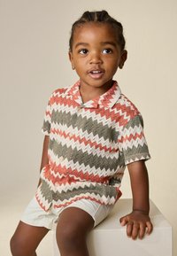 Junges Kind mit geflochtenem Haar, das ein gemustertes kurzärmliges Hemd und helle Shorts trägt, sitzt auf einem weißen Würfel vor einem einfarbigen Hintergrund.