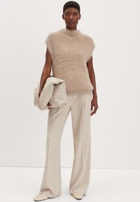 Top smagliante beige senza maniche con collo alto, abbinato a pantaloni a gamba larga beige chiaro. La modella tiene una borsa morbida di color crema.