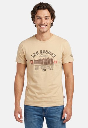 Homme souriant aux cheveux bruns ondulés portant un t-shirt beige Lee Cooper London Originals et un jean bleu, debout devant un fond blanc uni.