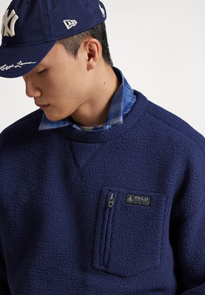 Giovane uomo che indossa una felpa in pile blu navy con tasca sul petto con cerniera e una camicia a quadri blu sotto, abbinata a un berretto da baseball blu.