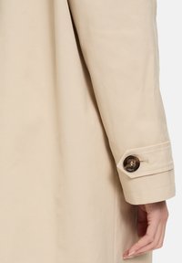 Manica di un trench coat beige chiaro con chiusura a bottone con finitura lucida e dettagli di cucitura sottili sul polsino.