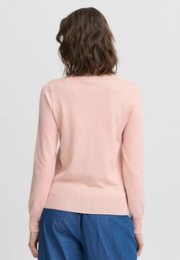 Maglione a maglia rosa chiaro con maniche lunghe, collo rotondo e orlo a costine. Texture liscia visibile, abbinato a pantaloni in denim blu.