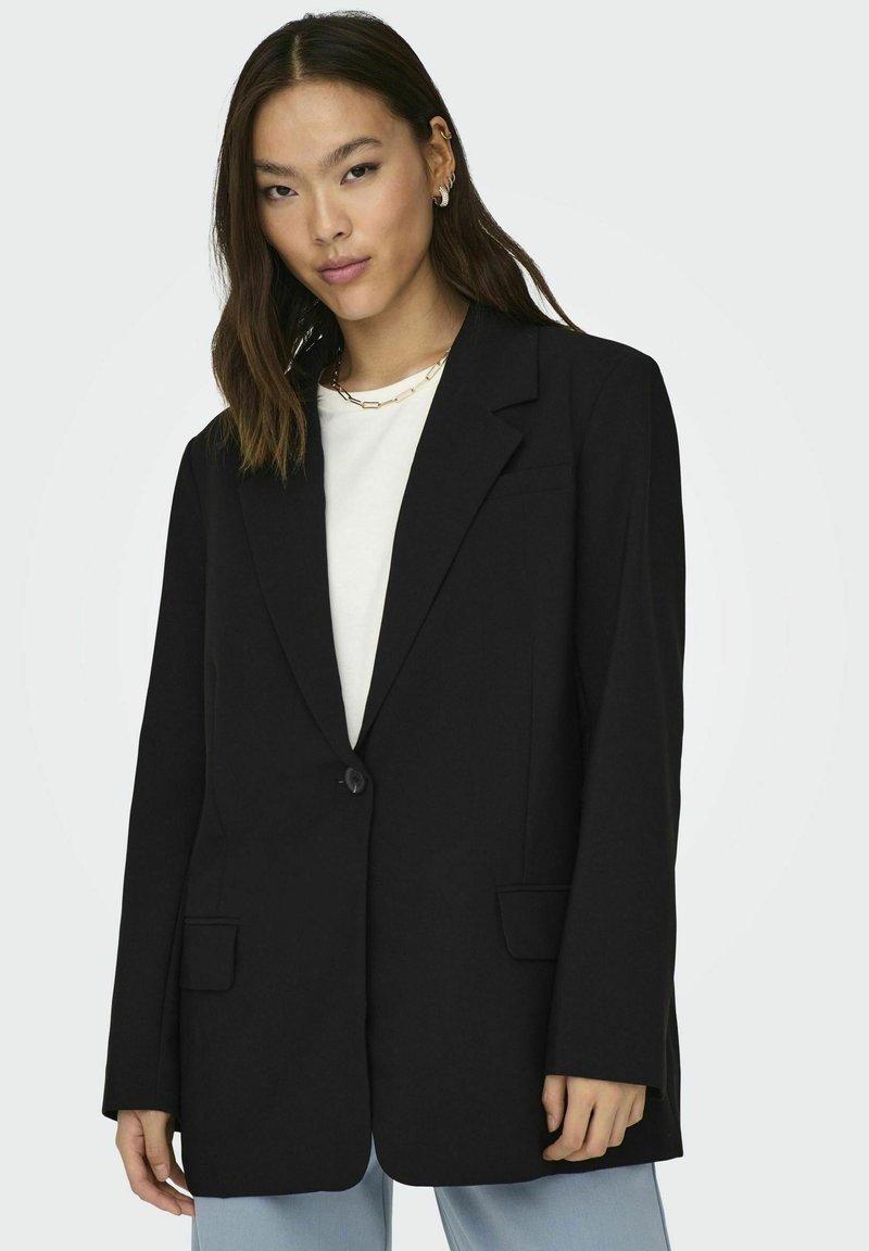 Schwarze maßgeschneiderte Blazer mit einem einzelnen Knopf, Reverskragen, zwei vorderen Taschen und strukturierter Form, getragen über einem weißen Oberteil.