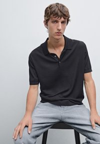 Černé polo tričko s krátkým rukávem a tříknoflíkovou légou, měkká textura, nošeno s světle šedými džíny. Model sedí na židli na jednobarevném pozadí.