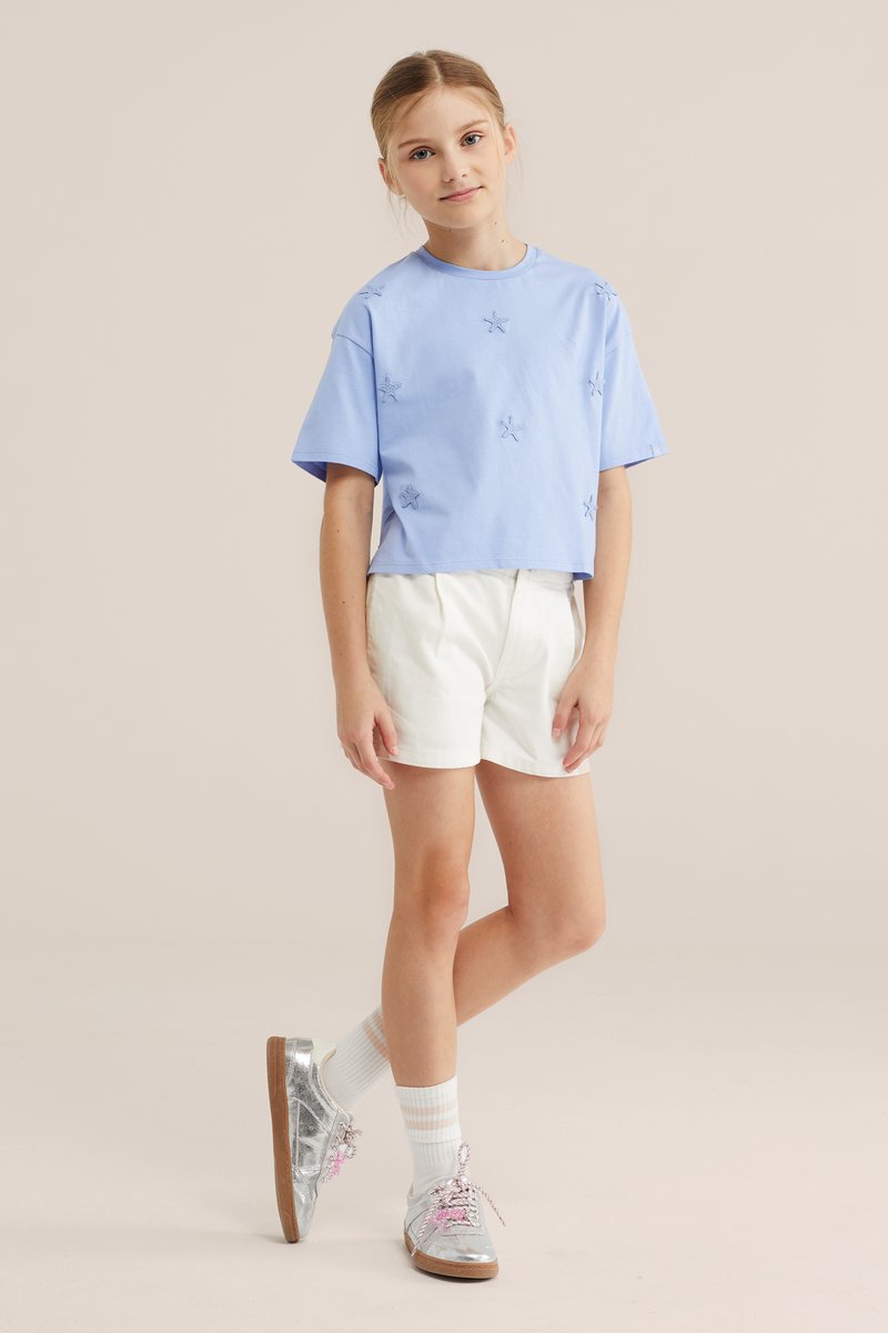 Jeune fille debout portant un t-shirt bleu clair orné d’étoiles, un short blanc, des chaussettes blanches et des baskets argentées brillantes avec des lacets roses.