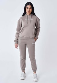 Hoody en bijpassende joggers van zacht, licht taupe stof. Voorzien van een voorzak en een geborduurd logo in het wit. Verstelbare koordjes.