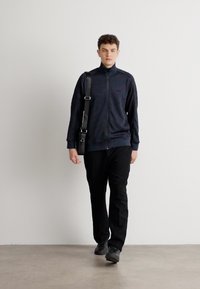 Námornícky zip-up sveter s vysokým golierom, čierne nohavice a čierne tenisky. Model nesie čiernu popruhovú tašku a kráča po hladkej podlahe.