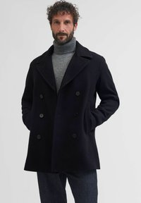 Melna vilnas peacoat ar dubulto pogu priekšpusi, lieliem jaciņu uzdariem un sānu kabatām, uzvilkta virs pelēka turtleneck. Gluda tekstūra un precīza piegriezuma.