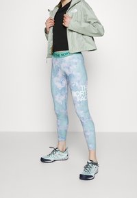 Ljusgrön vindjacka över en svart topp, ihop med pastellfärgade tie-dye leggings med "THE NORTH FACE"-logotyp och ljusblå vandringsskor.