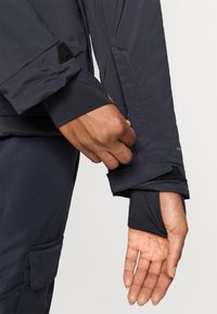 Jachetă neagră cu închidere Velcro la manșetă. Mâneca interioară, moale și elastică, este vizibilă, cu un material exterior neted și elegant.