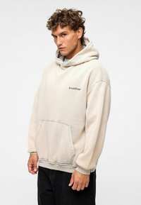 Beige hoodie met een voorzak en geribde mouwen, voorzien van een klein, donker geborduurd logo op de linkerborst.