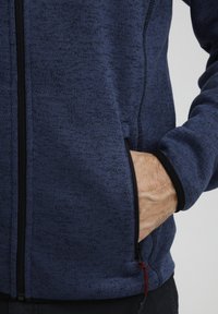 Blau strukturierte Zip-Jacke mit schwarzen Akzenten und Taschen. Verfügt über einen Kordelzug am Saum zur Anpassung. Weicher Stoff für Komfort.