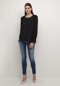 Juoda ilgomis rankovėmis bluzonė su nėrinių detalėmis prie kaklo, derinama su siaurais mėlynais džinsais ir sidabro spalvos smailiais bateliais.