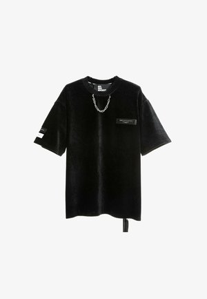 Camisa negra de gran tamaño hecha de pana, con mangas cortas, un acento de cadena y un bolsillo frontal con una etiqueta rectangular.