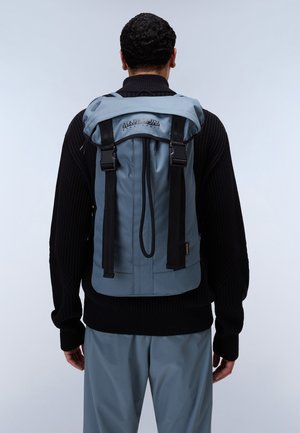 Rucksack aus hellblauem Stoff mit schwarzen Riemen. Verfügt über einen Rollverschluss, ein Logo auf der Vorderseite und verstellbare Seitengurte.