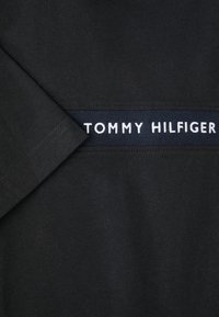 Crna pamučna majica s trakom od tamnoplave tkanine i bijelog vezanog logotipa "TOMMY HILFIGER", kratkih rukava s vidljivim šavovima.