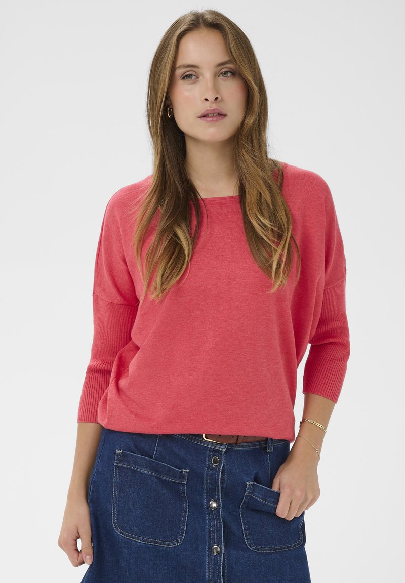 Pull en maille corail à la coupe décontractée et manches trois-quarts, associé à une jupe en jean foncé avec deux grandes poches avant et des boutons.