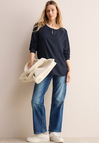 Marineblauw langemouwtop met een keyhole halssluiting, gecombineerd met lichtblauwe rechte jeans en witte sneakers, terwijl ze een crèmekleurige deken vasthoudt.