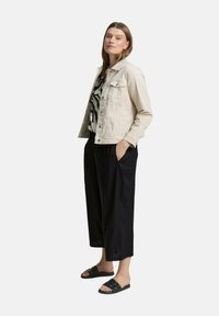 Chaqueta de mezclilla beige sobre una blusa de patrón blanco y negro, combinada con unos culottes negros sueltos y chanclas negras, de pie de forma casual.