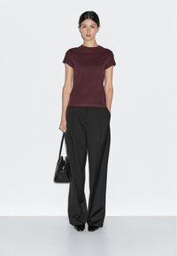 Burgundské tričko s krátkým rukávem v kombinaci s černými širokými kalhotami a černou kabelkou. Hladká látka, jednoduchý design a neutrální pozadí.