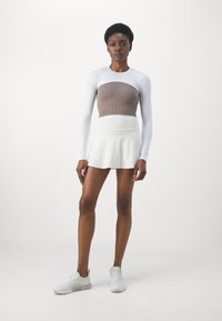 Dugi rukav atletska majica s prilagođenim smeškastim rebrastim korzetom, u paru s lepršavom belom suknjom. Model nosi bele tenisice. Minimalistički dizajn.